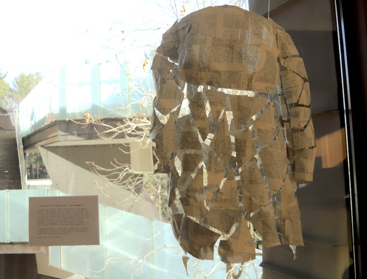 A body form of a young child in a dress, made out of paper mache book pages, fragmented but suspended together by string. The piece hangs vertically against a window.
